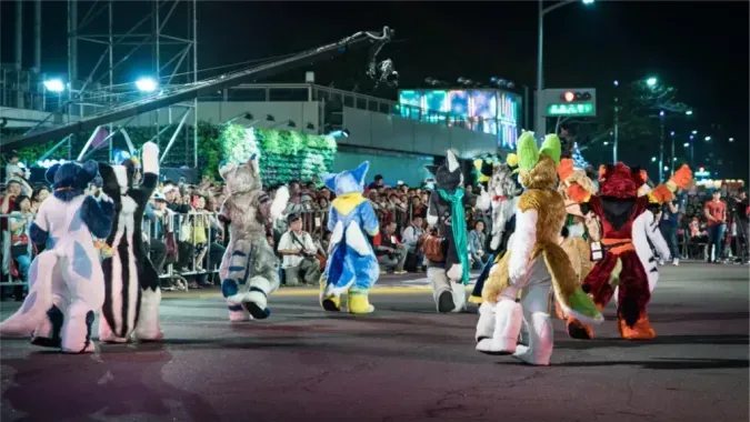During a nighttime parade, several fursuiters dance along the street as crowds watch from both sides.