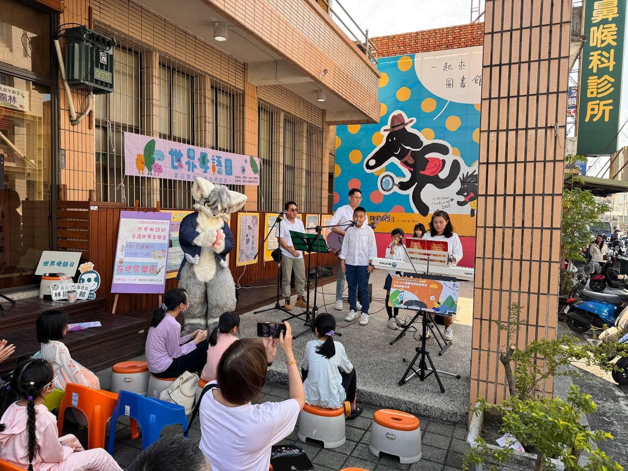 獸裝參加者站在林園分館前廣場，陪同兒童樂團演唱世界母語日活動節目，民眾席地觀賞。