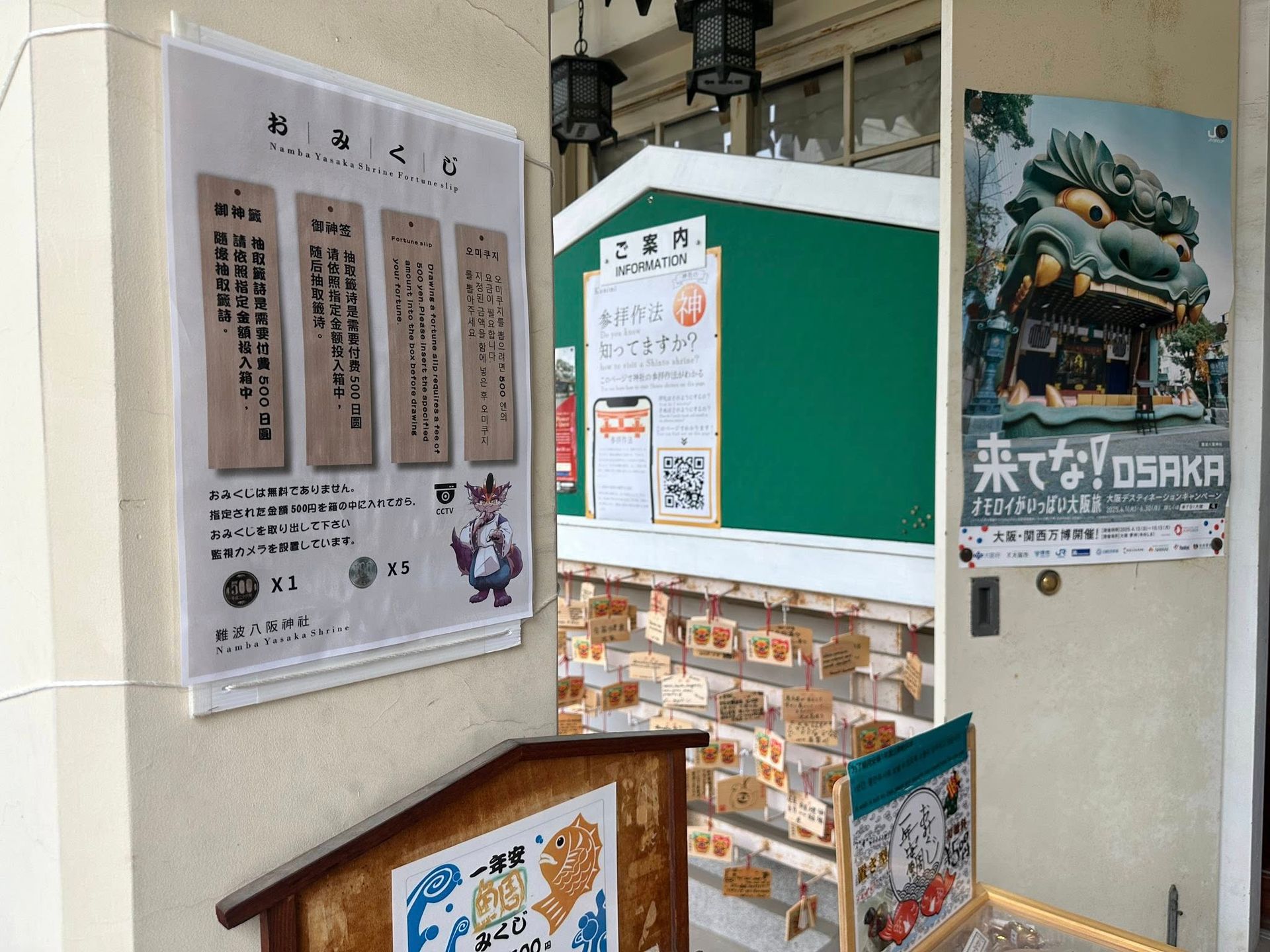 難波八阪神社入口的多語言御神籤說明看板,標示抽籤費500日圓,印有以茶會毛吉祥物插畫,一旁貼有參拜作法告示、QR code與大阪觀光宣傳海報。