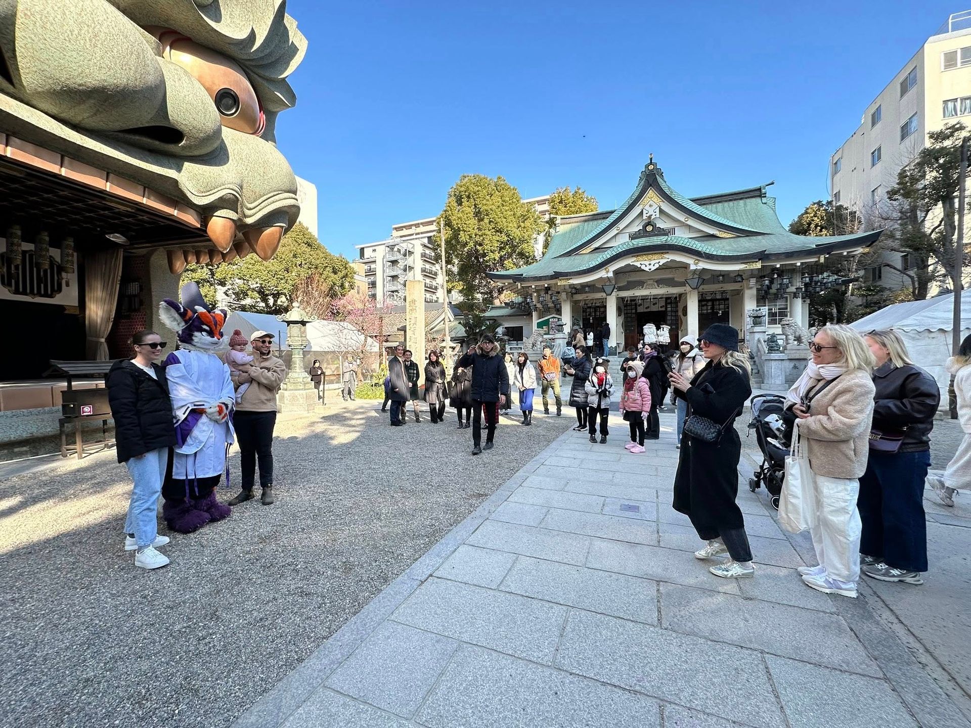 紫橙配色獸裝角色與遊客們站在難波八阪神社廣場,背景為巨大獅子頭造型的舞台裝置與神社本殿