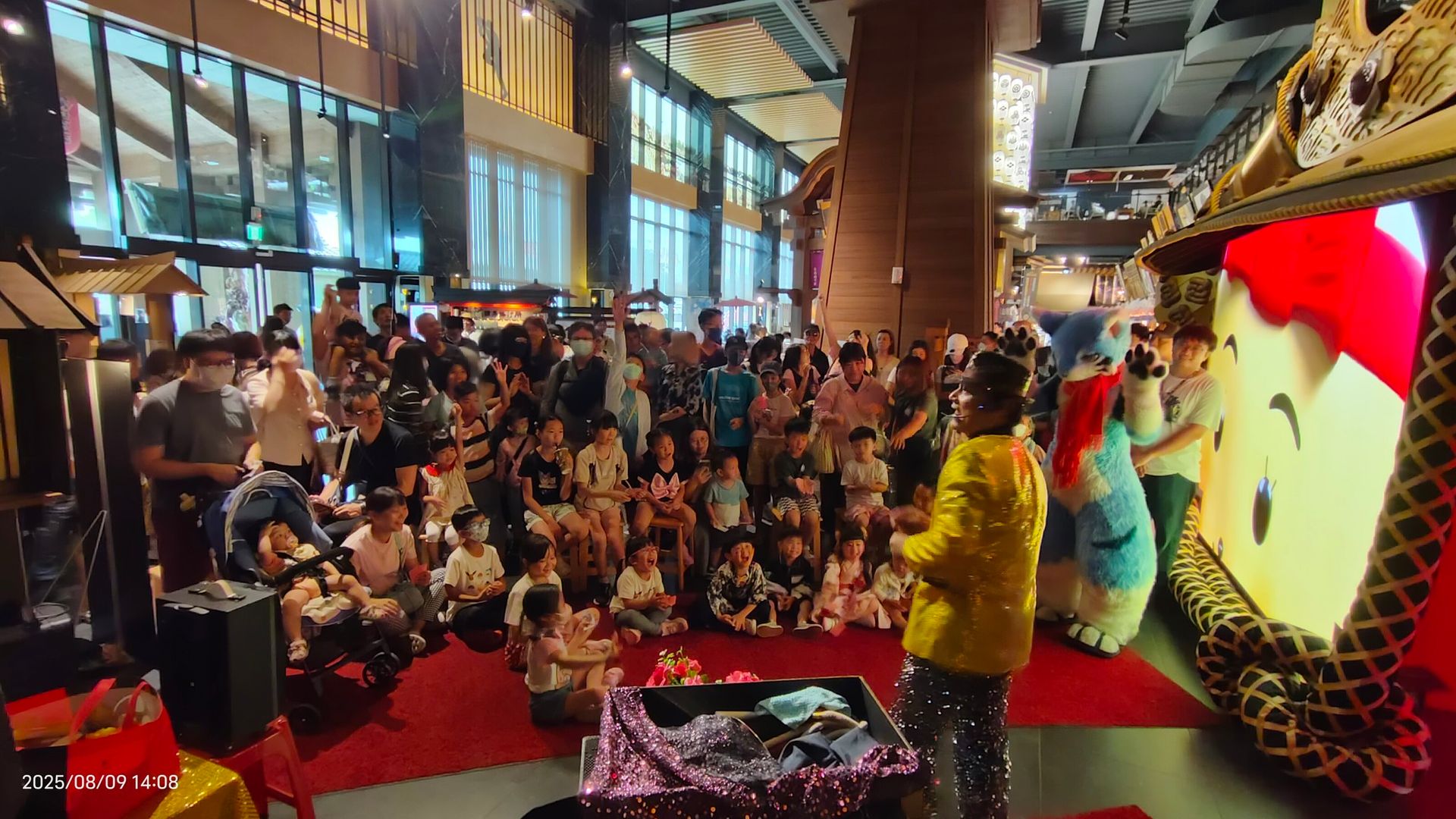 主持人在紅毯前對台下聚集的大批孩童與家長表演魔術，一旁站著藍色獸裝角色與大型招牌裝飾