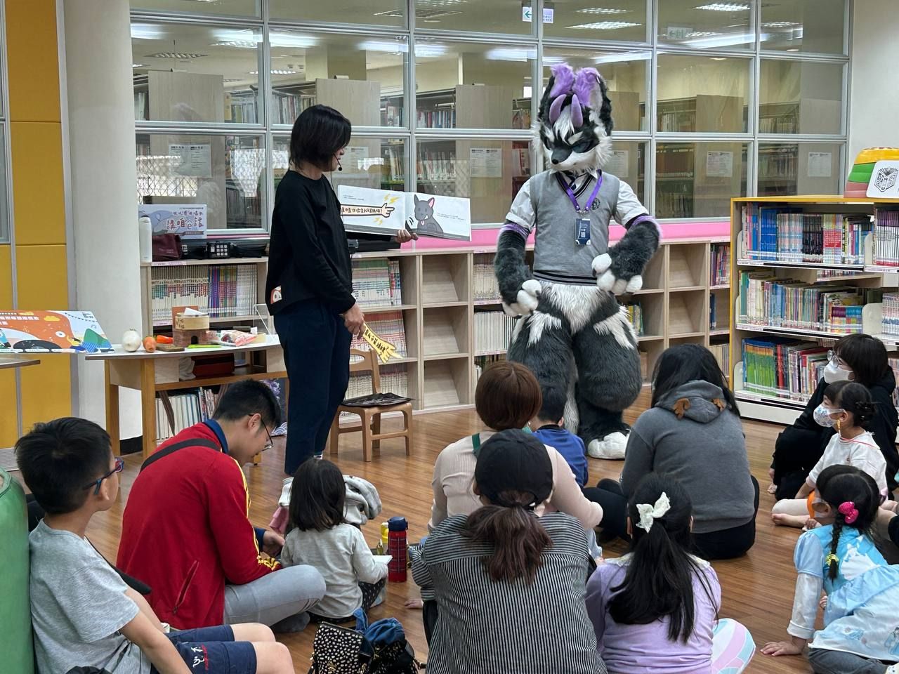 高雄市立圖書館大寮分館內，館員舉著繪本說故事，灰紫色狼型獸裝角色站在一旁，孩子們席地而坐專心聆聽。