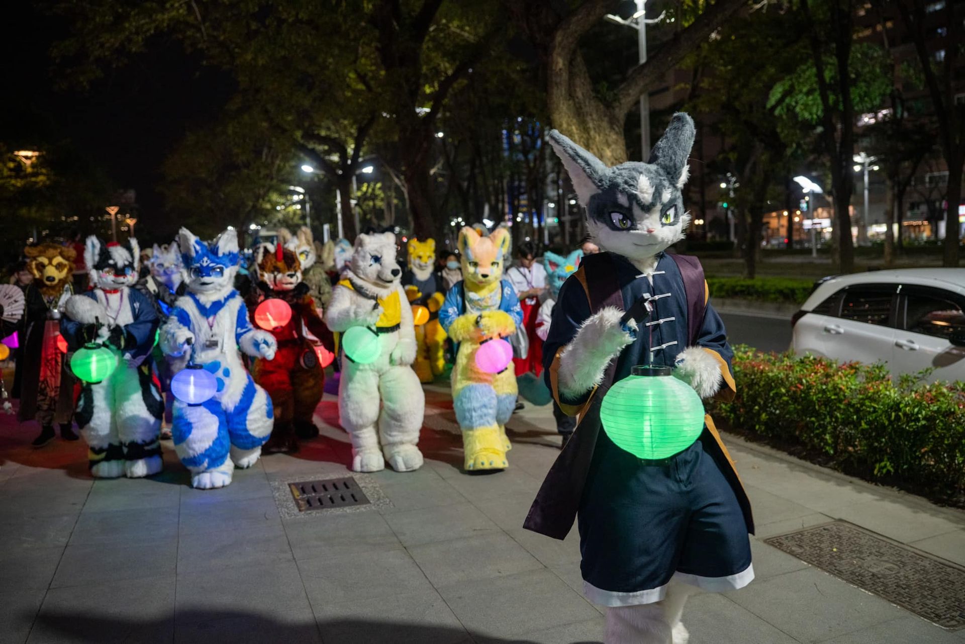 夜間提燈遊行隊伍走在市區人行道上，走在最前方的貓耳獸裝參加者手持綠色發光燈籠。