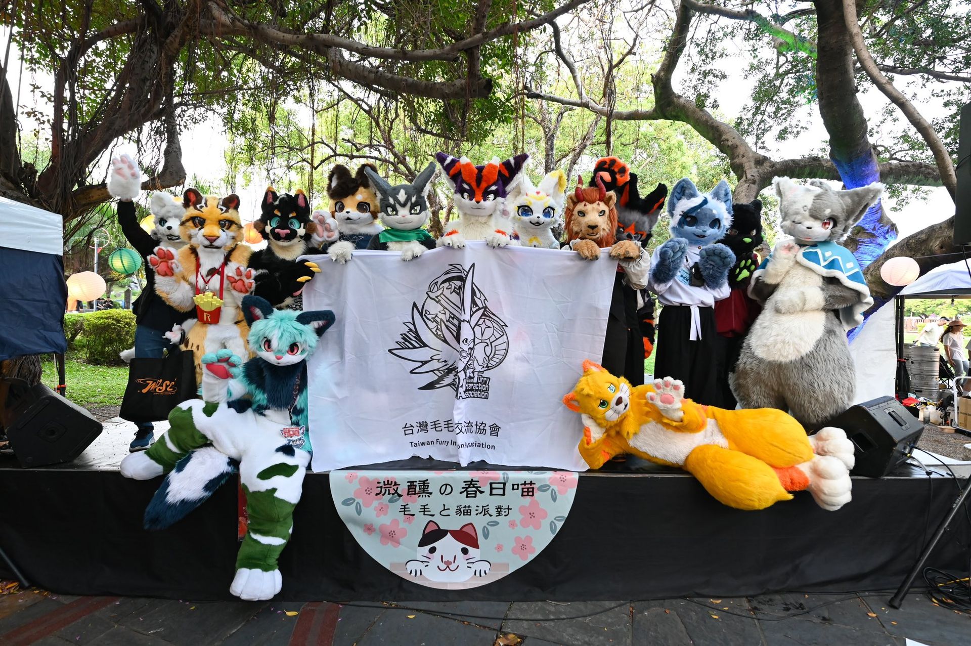 十多位獸裝參加者站在戶外舞台上合影，共同舉著印有台灣毛毛文化交流協會標誌與《微醺の春日喵》活動名稱的白色旗幟，背景為茂密老樹。