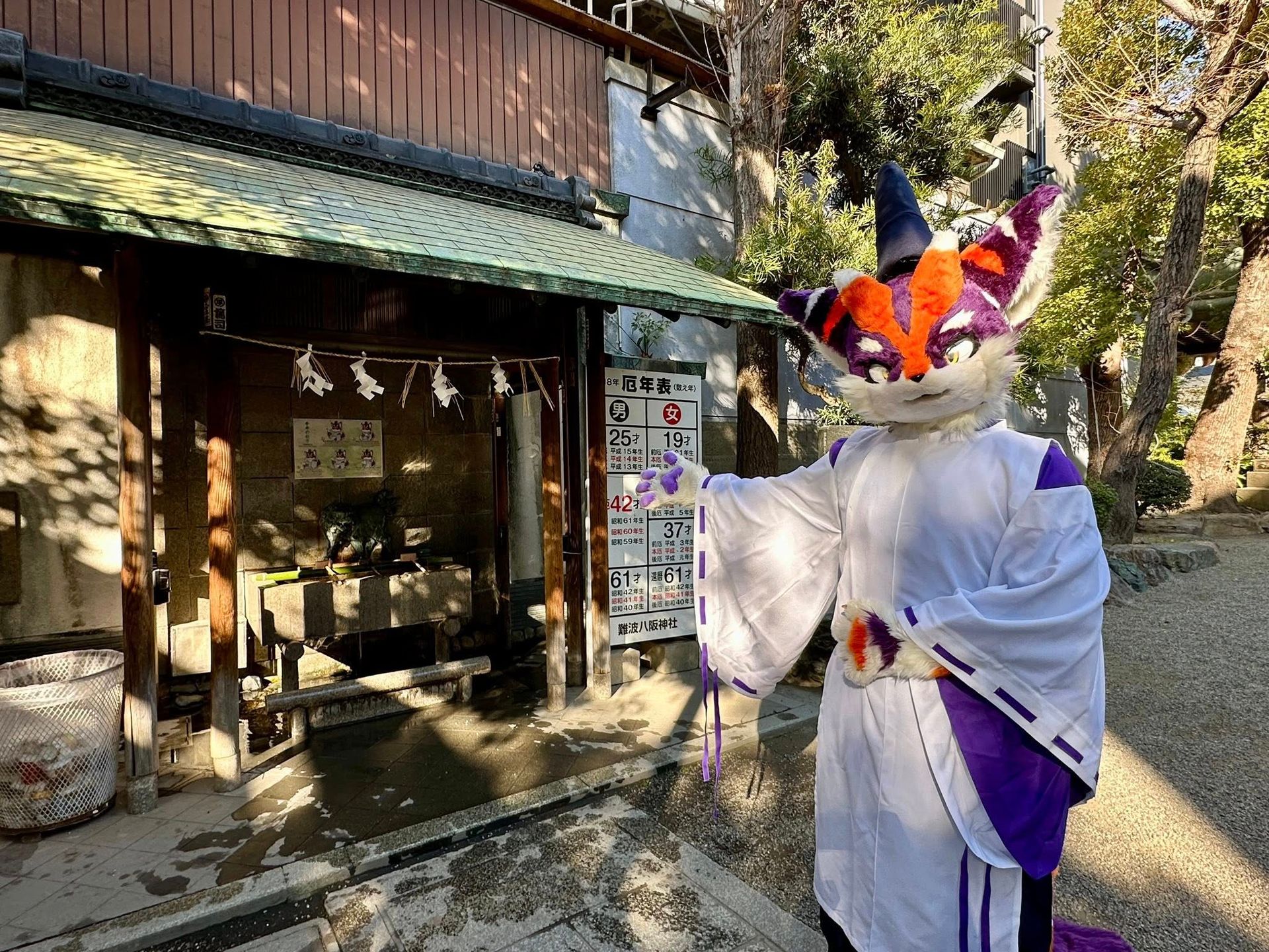 紫橙配色獸裝角色站在難波八阪神社的手水舍前,一旁立有厄年表告示牌