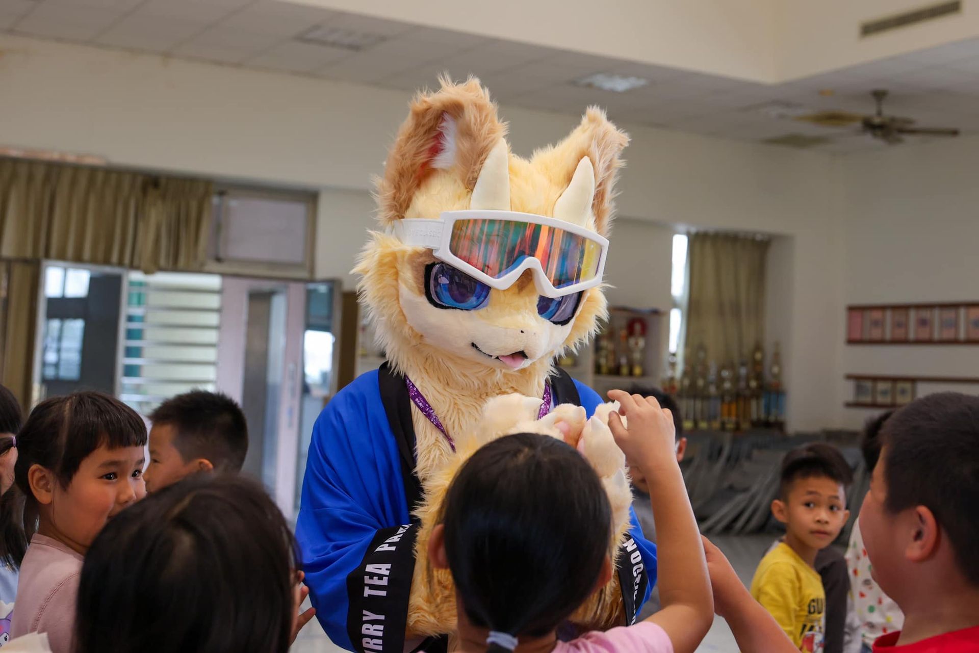橘黃色狐狸獸裝角色近距離與圍繞的孩子們互動，多名兒童伸手觸摸牠毛絨絨的手掌。