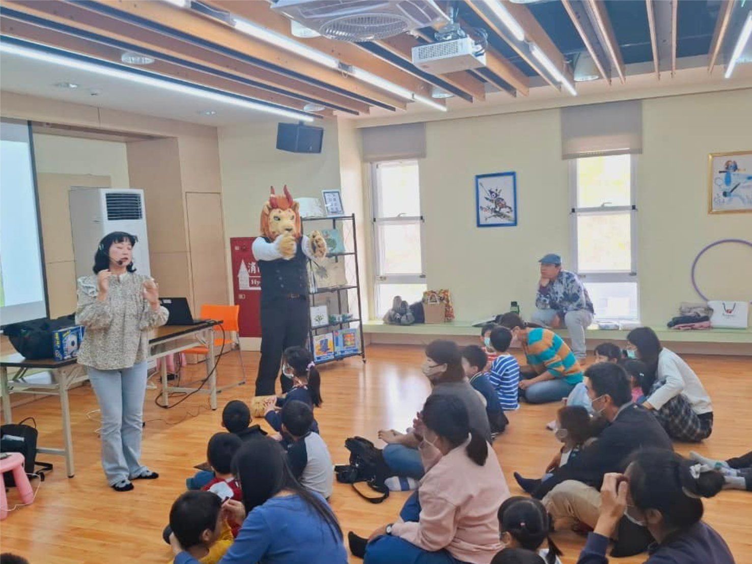 紅棕色獅子獸裝角色站在館員身旁帶領活動，孩子與家長席地而坐專心聆聽台語主題分享。