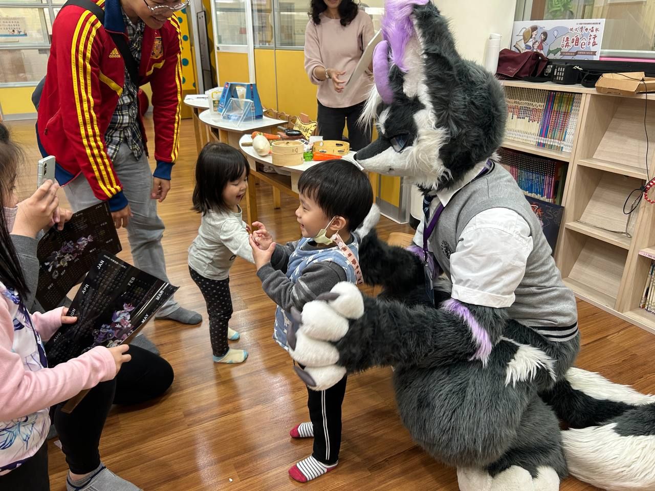 灰紫色狼型獸裝角色蹲下與兩名幼童擊掌互動，一旁家長拿著月曆式紀念小卡拍照留念。