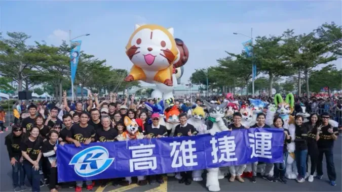 A large group of fursuiters and staff pose outdoors holding a "Kaohsiung MRT" banner, with a giant inflatable cat balloon behind them.