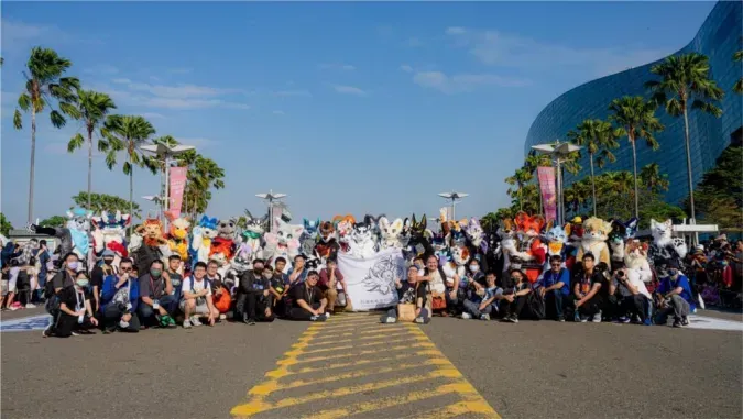 Fursuiters pose in a long row on the street outside Dream Mall, with someone holding up a white group banner in the middle.