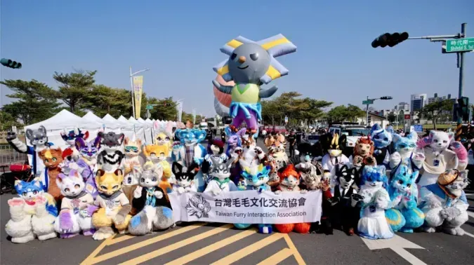 Fursuiters line up for a group photo on the street with a giant bird-shaped mascot balloon behind them, displaying the "Taiwan Furry Interaction Association" banner.