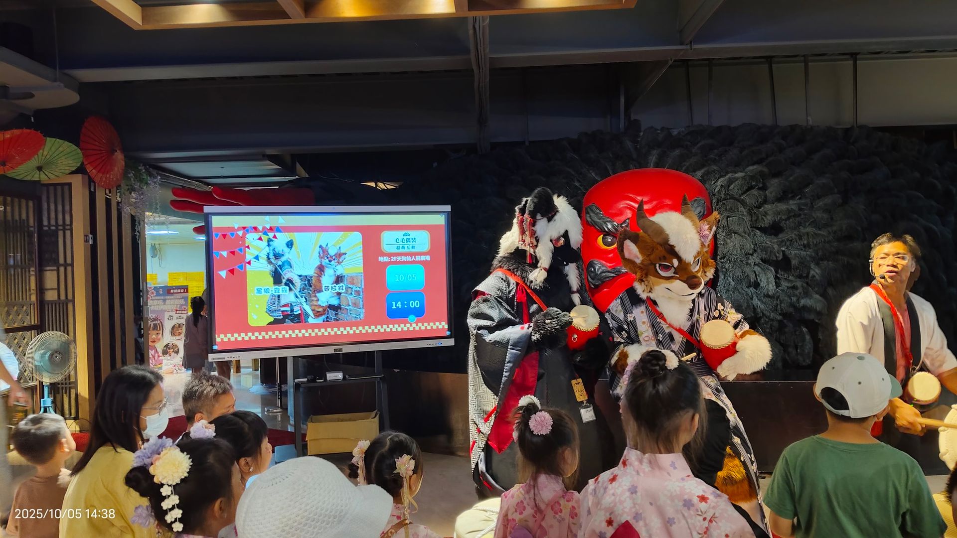 獸裝角色在室內舞台表演太鼓，背景螢幕顯示活動資訊與日期時間，台下坐著穿浴衣的孩童觀眾