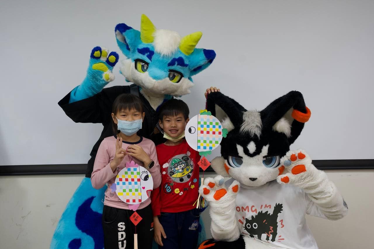 藍色龍型獸裝角色與黑白貓型獸裝角色和兩名小朋友合影，孩子們手持自製的彩繪魚形吊飾。
