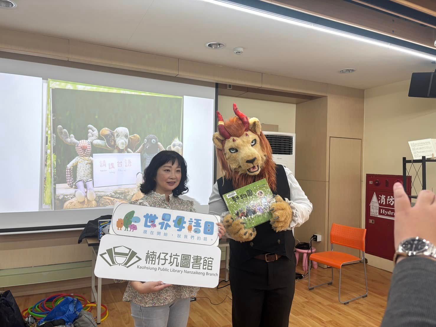 紅棕色獅子獸裝角色與館員一同展示「世界母語日」與「楠仔坑圖書館」立牌，手拿繪本《動物園》，背景投影台語玩偶教材畫面。