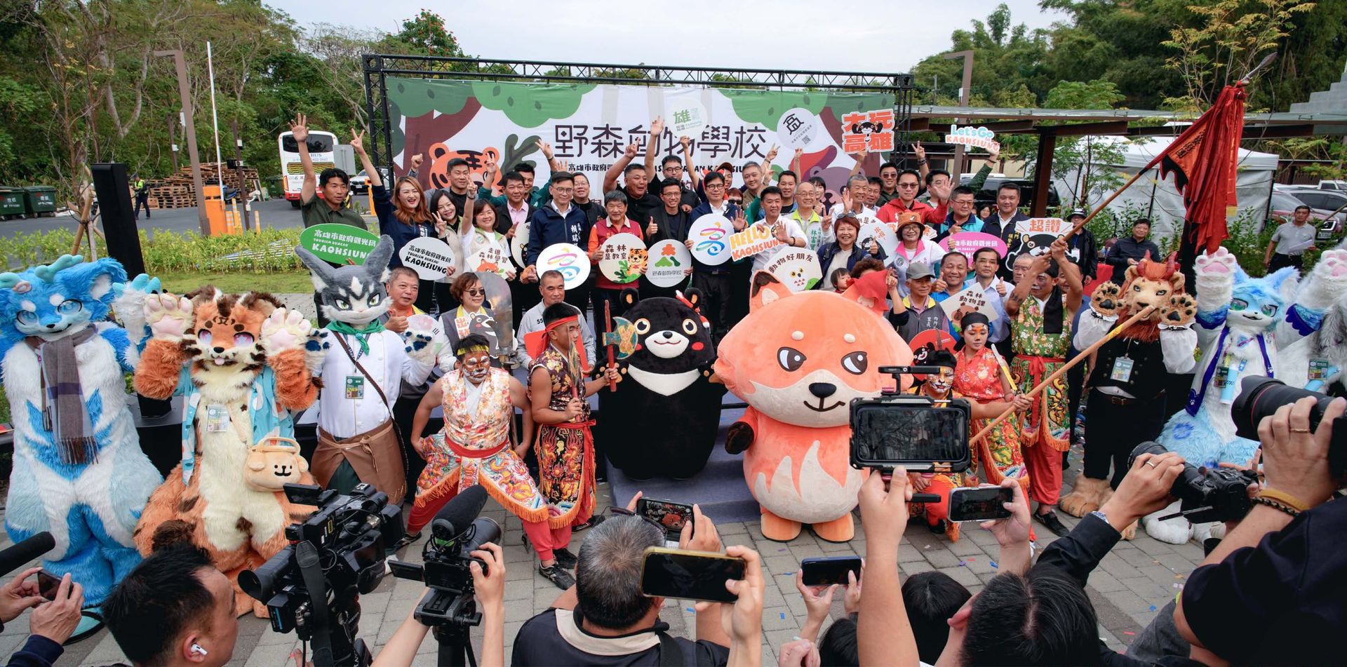 野森動物學校開幕活動大合照，獸裝參加者與市府官員、熊讚等吉祥物一同在台上擺姿勢，台下媒體正在拍攝
