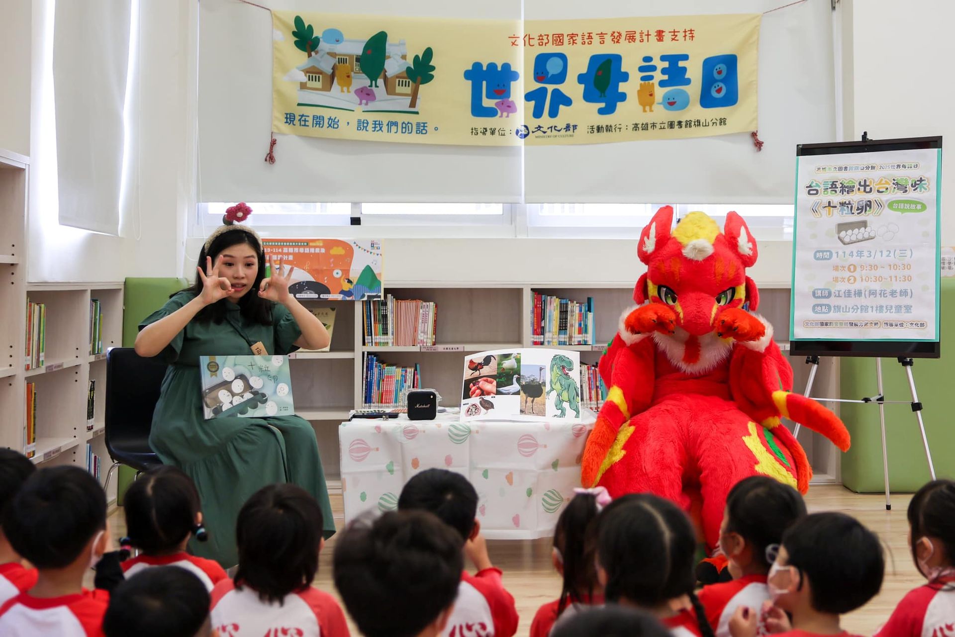 紅色龍型獸裝角色坐在館員身旁，一起以台語演出繪本《十粒卵》故事，背後掛著「世界母語日」布條，學童席地聆聽。