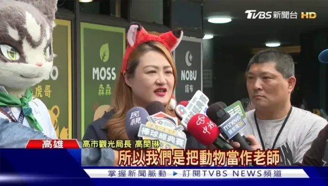A TVBS News still of Kaohsiung Tourism Bureau Director Kao Min-lin being interviewed, saying "So we treat animals as teachers," with a gray cat fursuiter visible at the left edge.