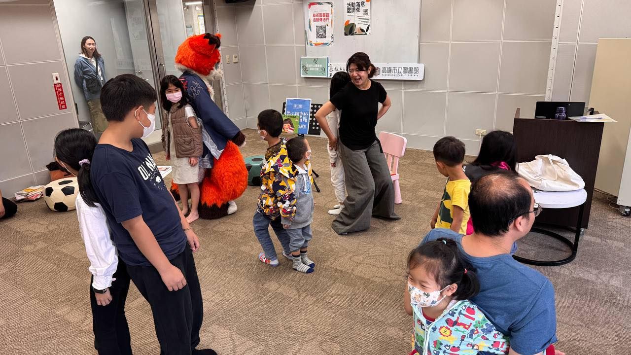 橘色狐狸獸裝角色站在高雄市立圖書館草衙分館活動室內,與多位小朋友及家長互動,牆上貼著活動滿意度調查看板。