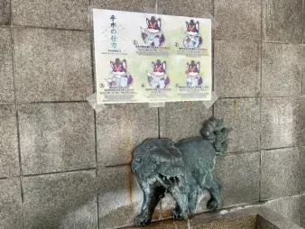 A shrine's water purification basin (temizuya), with the fox character's step-by-step guide posted above it, and an animal-shaped spout over the stone basin.